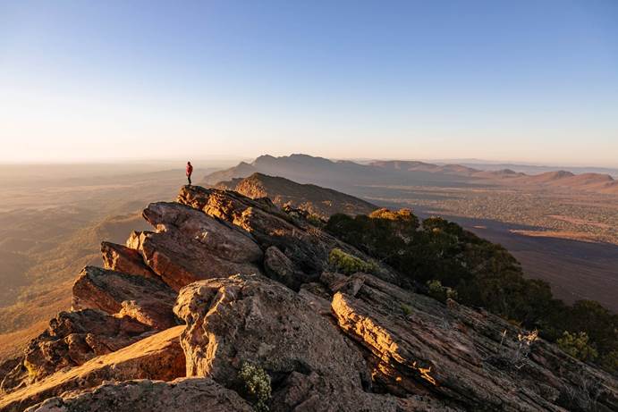 A woman stands on a mountain peak in an arid landscape.